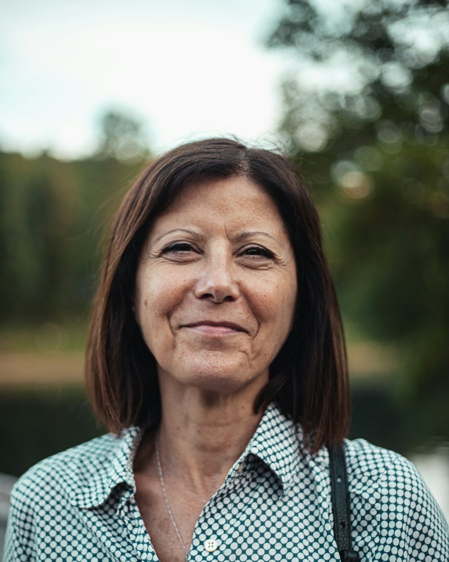 Portrait of a smiling mature woman with shoulder-length brown hair and a green-and-white patterned shirt, outdoors in soft natural light