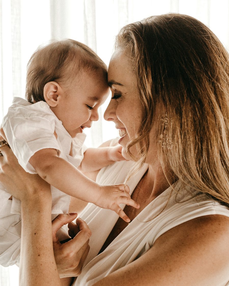 A smiling mother lifts her baby so their foreheads touch, in soft window light