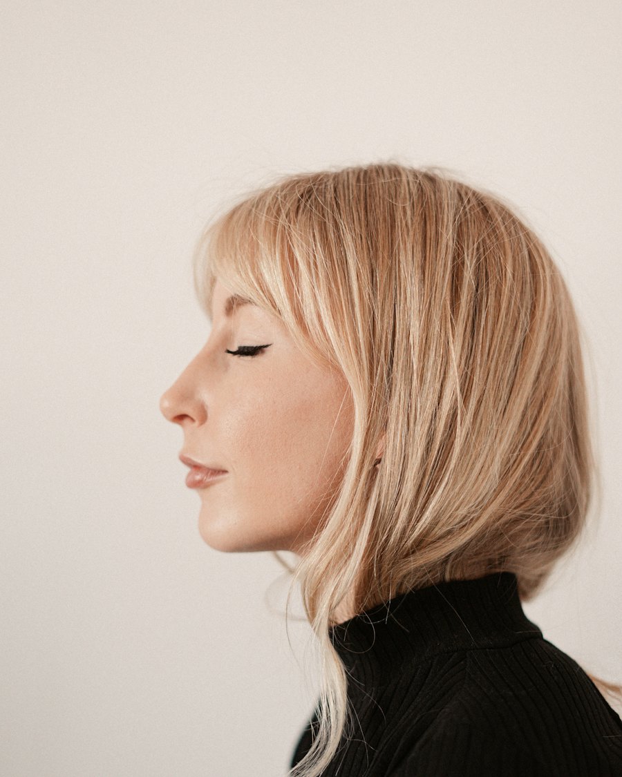Side profile of a woman with a blonde bob and closed eyes, wearing a black turtleneck, against a plain cream wall