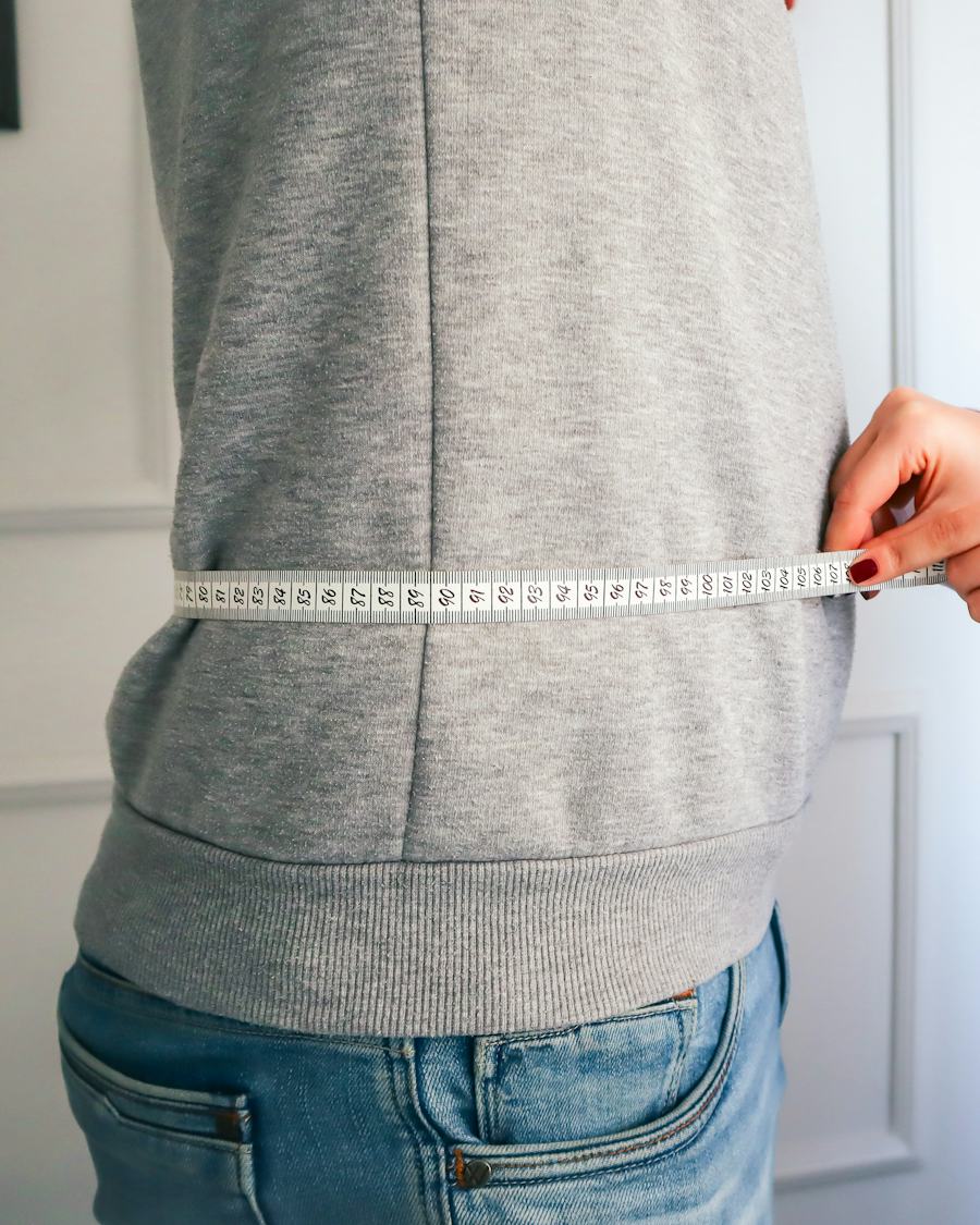 A hand wraps a measuring tape around a person's waist over a gray sweatshirt and jeans
